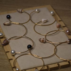 May include: Gold-toned metal bracelets, each adorned with a natural seashell or pearl. The bracelets are displayed on a wooden stand with a beige backing, showcasing a variety of designs and colors.