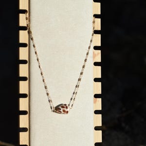 May include: A gold-coloured necklace featuring a small, patterned pendant. The pendant is brown and white, and the chain has a rectangular link design. The necklace is displayed against a light-coloured, textured backdrop.