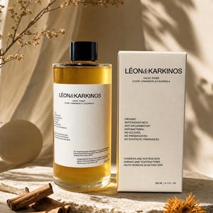 May include: A clear glass bottle and a white box of facial toner, with the brand name "L&Eacute;ON & KARKINOS". The bottle contains a golden liquid, and the box lists ingredients like clove, cinnamon, and calendula. Cinnamon sticks and cloves are arranged near the bottle.