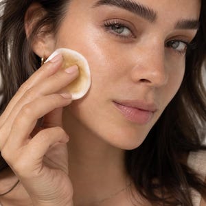 May include: A person holding a round, beige cotton pad to their cheek. The pad appears to be used for skincare. The person has brown hair, green eyes, and is wearing a gold necklace and earrings.
