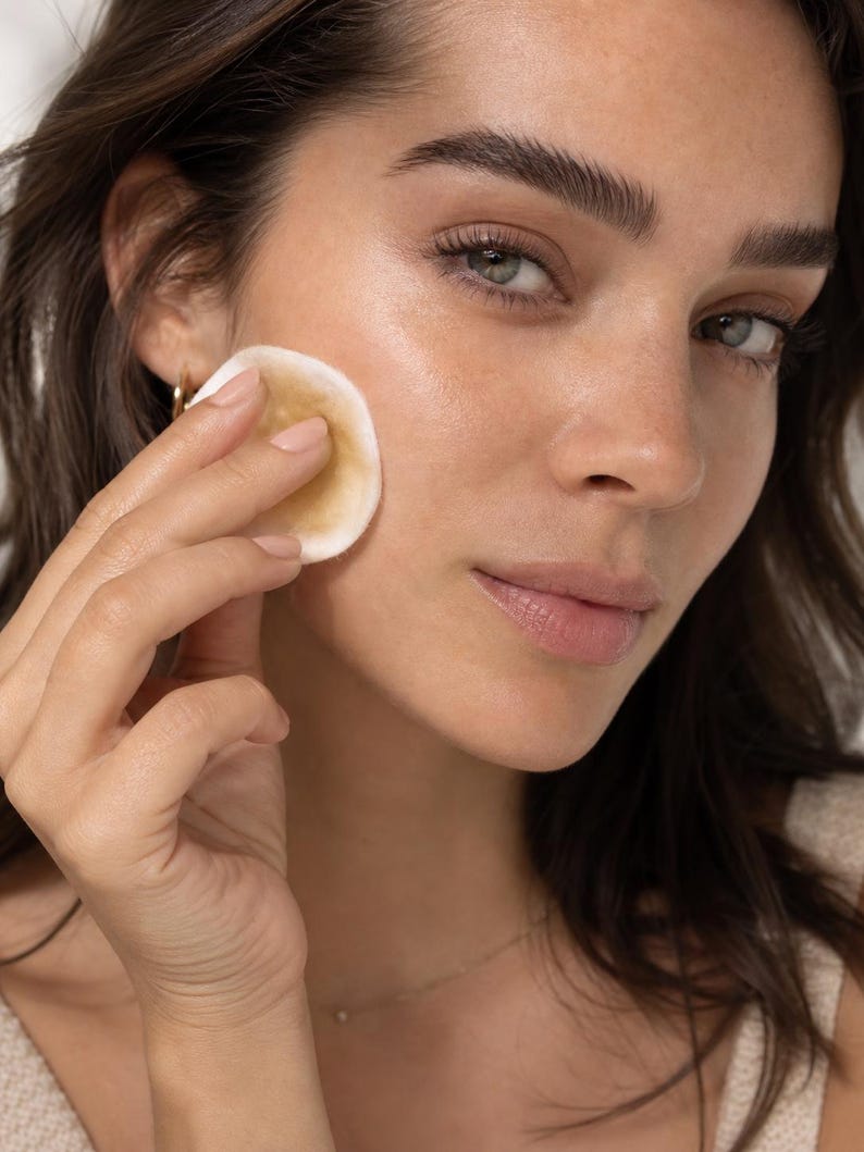 May include: A person holding a round, beige cotton pad to their cheek. The pad appears to be used for skincare. The person has brown hair, green eyes, and is wearing a gold necklace and earrings.