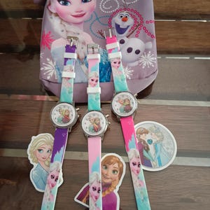 May include: Three children's watches with colourful straps featuring Elsa from Frozen. The watch faces show Elsa and Anna. A matching purple bag with Elsa and Olaf is in the background. Stickers of Elsa and Anna are also present.