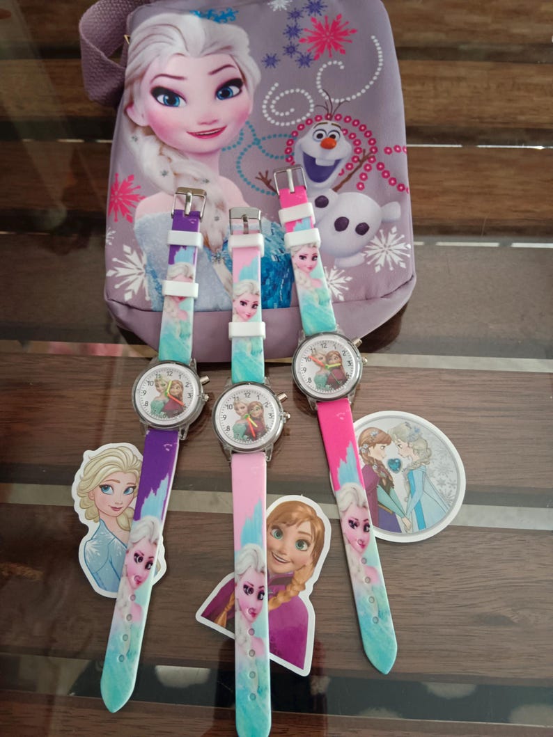 May include: Three children's watches with colourful straps featuring Elsa from Frozen. The watch faces show Elsa and Anna. A matching purple bag with Elsa and Olaf is in the background. Stickers of Elsa and Anna are also present.