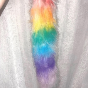 May include: A long, plush faux fur tail featuring a vibrant rainbow gradient. The colors transition vertically from a soft pink at the top, through orange, yellow, green, and blue, ending in a light purple and white tip with a thick, shaggy texture.