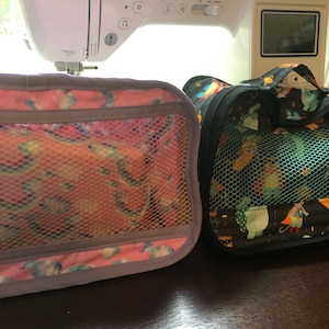 May include: Two zippered travel bags with mesh panels. The bag on the left is purple with a pink and white floral pattern. The bag on the right is black with a colorful cartoon animal print.