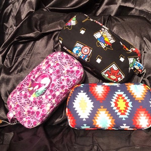 May include: Three different patterned zippered pouches. One pouch is black with a white and yellow geometric pattern and features cartoon characters. Another pouch is purple with a floral pattern. The third pouch is blue with a red, white, and yellow geometric pattern.