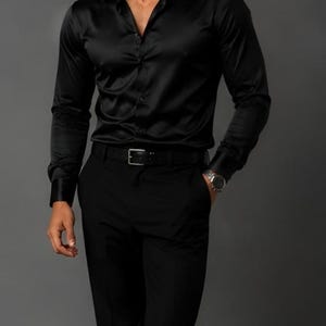 May include: A long-sleeved black button-down shirt with a smooth, satin-like finish, paired with matching black trousers and a black leather belt. The shirt features a classic collar, buttoned cuffs, and a front button placket, creating a monochromatic look.