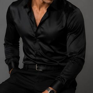 May include: A long-sleeved black button-down shirt with a smooth, satin-like finish, featuring a classic collar, buttoned cuffs, and a front button placket. The shirt is paired with black trousers, a black leather belt with a silver buckle, and a watch.