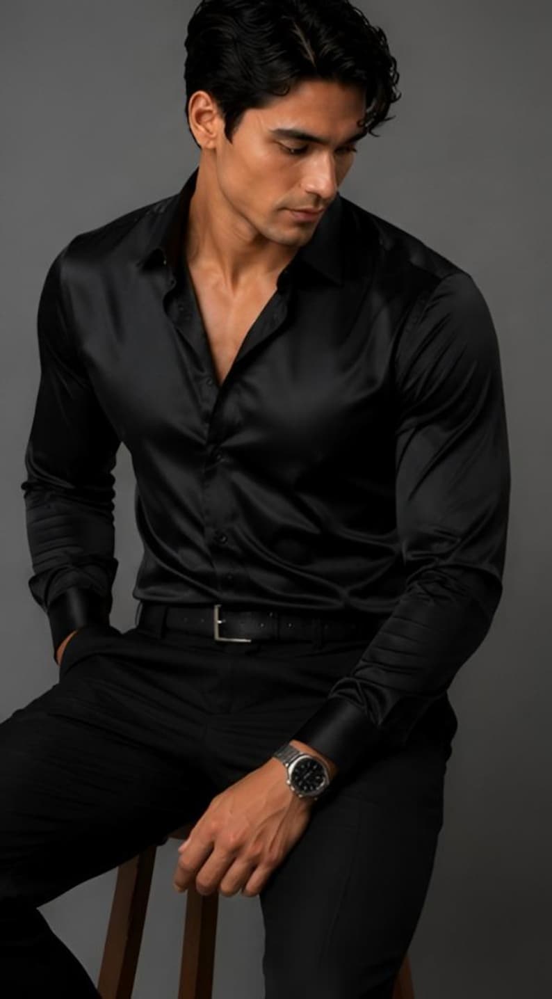 May include: A long-sleeved black button-down shirt with a smooth, satin-like finish, featuring a classic collar, buttoned cuffs, and a front button placket. The shirt is paired with black trousers, a black leather belt with a silver buckle, and a watch.