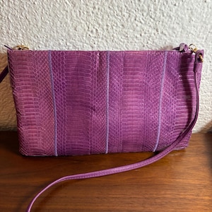 May include: A rectangular purple crossbody bag featuring a textured snakeskin pattern. The bag is constructed from vertical panels of varying shades of purple, accented by thin, lighter purple piping. It includes a thin, matching purple shoulder strap.