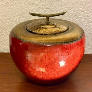 May include: A round ceramic jar with a vibrant red glazed body and a textured, metallic bronze-toned lid. The lid features a central, horizontal T-shaped handle with a weathered finish, contrasting with the smooth, glossy red surface of the jar.