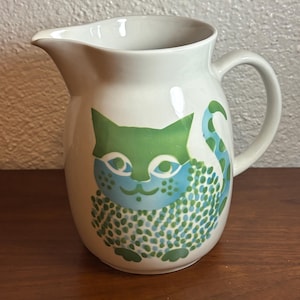 May include: A white ceramic pitcher featuring a stylized cat illustration on the front. The cat is depicted with a green head, a blue face, and a body covered in a pattern of small green and blue dots. The pitcher has a curved handle and a spout.