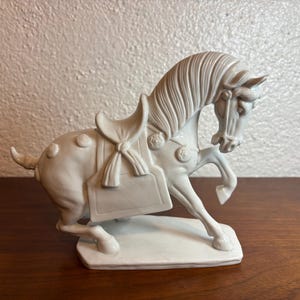 May include: A white ceramic horse figurine in a prancing pose, featuring a detailed saddle with a tied bow, a bridle, and a flowing mane. The horse is sculpted with circular decorative accents on its hindquarters and chest, standing on a base.