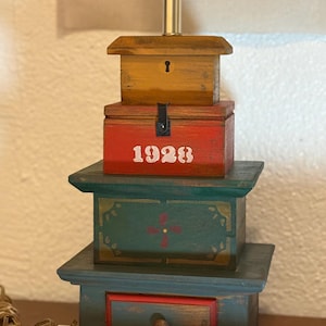 Puede incluir: Una base de lámpara de mesa construida con cajas de madera apiladas. La caja superior es de color marrón claro con una cerradura, la del medio es roja con el texto blanco 1928, y las dos inferiores son verdes con motivos florales pintados.