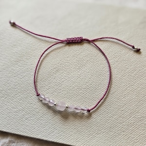 May include: A delicate bracelet featuring light pink rose quartz beads strung on a deep purple cord. The adjustable bracelet has a sliding knot closure and small silver-coloured accents. The bracelet is displayed on a textured, off-white surface.