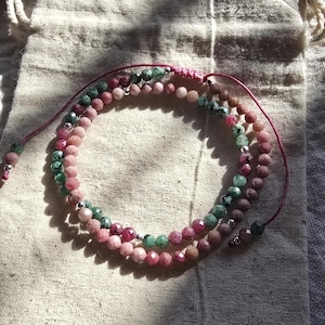 May include: A multi-strand beaded bracelet featuring faceted round beads in shades of pink, rose, and green. The bracelet is strung on a thin, dark pink cord with adjustable sliding knots and small silver-toned metal spacer beads between the stones.