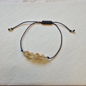 Könnte beinhalten: Verstellbares Armband mit vier hellgelb-braunen Edelsteinperlen. Die Perlen sind auf einer dunkelgrauen Kordel mit einem Schiebeknotenverschluss aufgefädelt. Kleine silberfarbene Perlen befinden sich zwischen den Edelsteinen und an den Enden der Kordel.