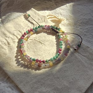 May include: A multi-strand bracelet featuring small, faceted, multicoloured gemstone beads in shades of pink, green, yellow, and clear. The bracelet includes small silver-toned metal spacers and a dark brown adjustable cord with two dangling beads.