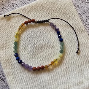 May include: A bracelet featuring a series of small, faceted, multi-colored gemstone beads arranged in a circular pattern. The beads include shades of red, yellow, green, blue, and purple. It has a thin black cord with an adjustable sliding knot.