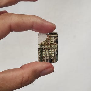 Könnte beinhalten: Ein kleiner rechteckiger Anhänger mit einer detaillierten handgemalten Illustration einer historischen Gebäudefassade mit Bogenfenstern und architektonischen Säulen in Braun-, Grün- und Weißtönen, mit einem kleinen Loch an der Oberseite.