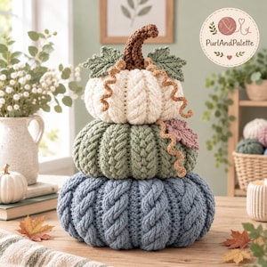 May include: A stacked crochet pumpkin decoration featuring three tiers in cream, sage green, and dusty blue. Each pumpkin has a cable-knit texture, a brown stem, and decorative green leaves and curly tendrils. The top pumpkin features the text PurlAndPalette.
