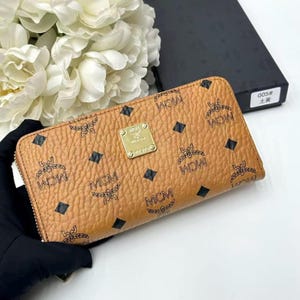 May include: A tan leather wallet with a zip closure, featuring a repeating pattern of the letters "MCM" and black diamond shapes. A gold-colored metal plaque is centered on the wallet. The wallet is displayed with white flowers and a black box in the background.