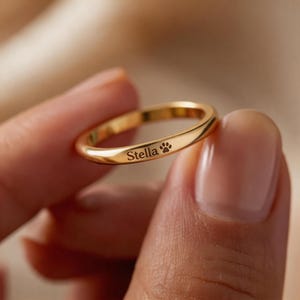 Può includere: Un anello a fascia sottile color oro con una finitura liscia e lucida. La superficie esterna dell'anello in metallo è incisa con il nome Stella seguito da una piccola icona a forma di impronta di zampa, creando un design semplice per questo gioiello.