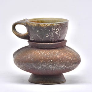 May include: A handcrafted ceramic cup and vase set. The cup is dark brown with a handle and decorative etchings. The vase is a bulbous shape with a textured surface and a dark brown and reddish-brown color scheme. The set is on a white background.