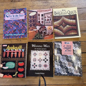 May include: Six instructional books on quilting arranged in two rows. Titles include Fit To Be Tied, Companions Quilts and Miniatures, Bargello Quilts, Appliquilt, Winning Ways To Make Quilts, and On to Square Two, featuring various quilt patterns and designs.