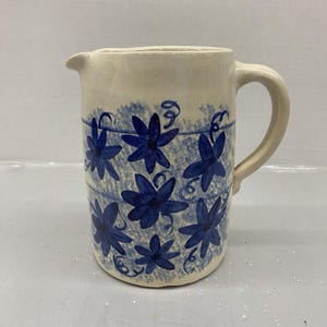 May include: A cream-colored ceramic pitcher featuring a hand-painted pattern of dark blue star-shaped flowers and swirling vine accents. The design is set against a textured blue sponge-painted background, complete with a sturdy handle and a spout.