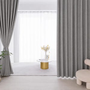 Puede incluir: Cortinas grises de suelo a techo con un acabado de tela texturizada, que presentan pliegues verticales profundos que cuelgan desde la parte superior. Las cortinas enmarcan una ventana luminosa con visillos blancos, una pequeña mesa cilíndrica dorada y un florero.