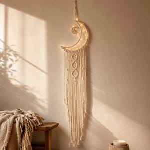 Op de afbeelding: Een wandhanger in de vorm van een halve maan gemaakt van off-white macramékoord, voorzien van een kleinere maanuitsparing en geïntegreerde warmwitte LED-verlichting. Het ontwerp bevat ingewikkelde knooppatronen en lange, loshangende franjes.