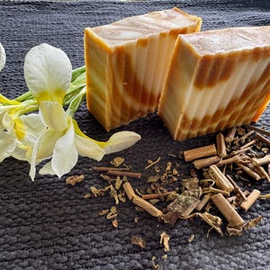 May include: Two bars of handmade soap with a layered, brown and white design. White and yellow iris flowers and dried herbs are arranged on a dark gray textured surface. The soap appears to be natural and organic.
