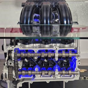 May include: A detailed display of a car engine, showcasing its internal components. The engine is illuminated with blue lights, highlighting the intricate design. The top section features a black intake manifold. The engine is mounted on a clear glass surface.