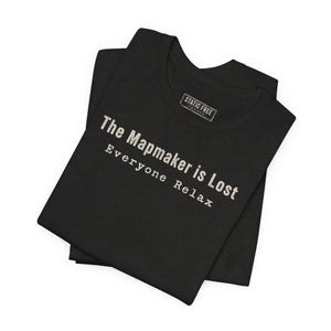 May include: A black t-shirt featuring the white text "The Mapmaker is Lost" and "Everyone Relax" printed on the front. The shirt includes a rectangular inner neck label with the text "STATIC FREE" and additional small printed details.