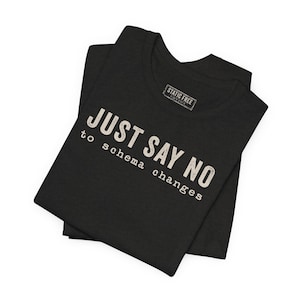 May include: A black short-sleeved t-shirt featuring the white text "JUST SAY NO to schema changes" printed on the front. The shirt includes a rectangular neck label with the text "STATIC FREE" visible near the collar area of the garment.