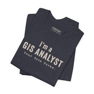 May include: A dark heather charcoal gray short-sleeve t-shirt featuring the white text "I'm a GIS ANALYST Your Data Sucks." printed on the front. The shirt includes a rectangular "STATIC FREE" brand label on the inside of the collar area.