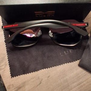 May include: Black sunglasses with a black frame and dark lenses, resting on a black cloth. The sunglasses are in front of a black box with the text "SEE BETTER WORLD" and the brand name "LUENX".