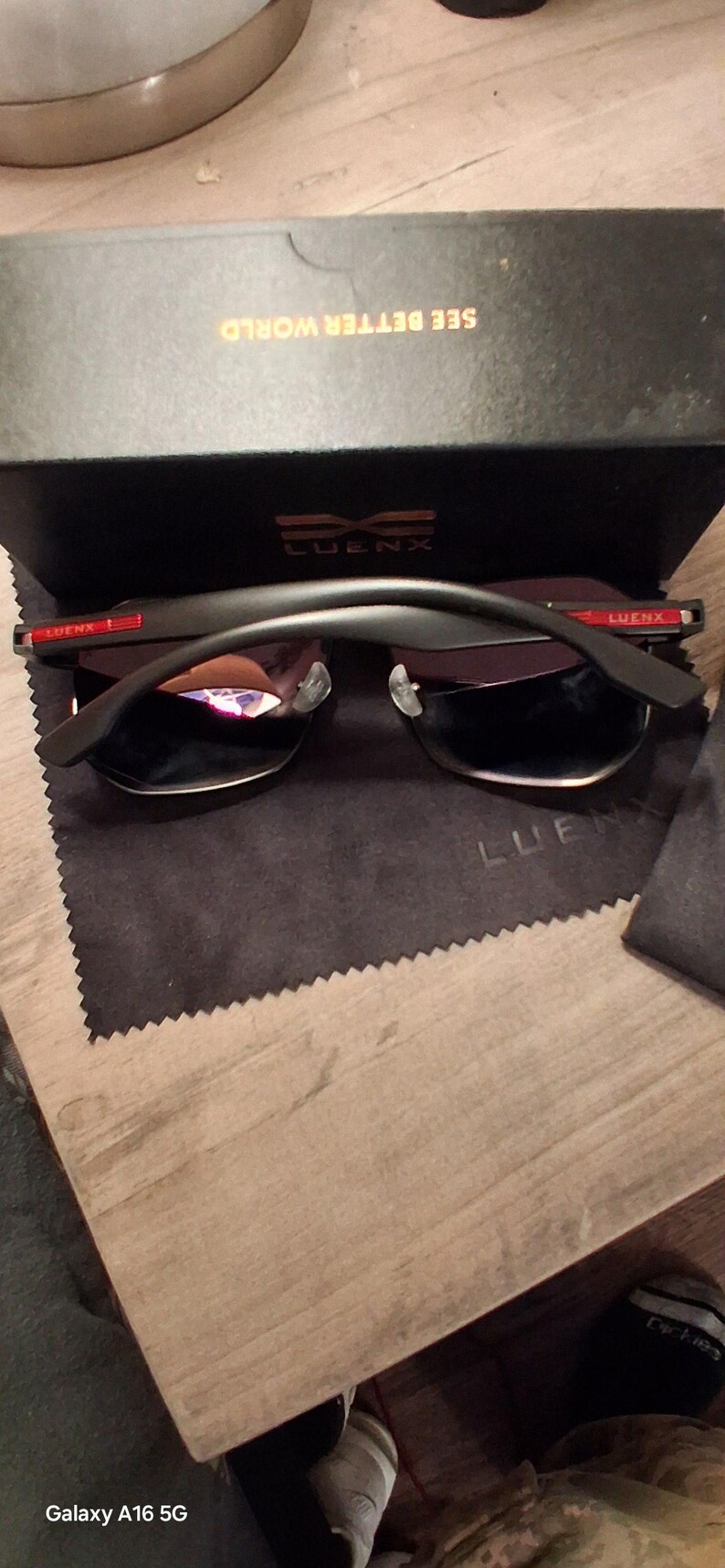 May include: Black sunglasses with a black frame and dark lenses, resting on a black cloth. The sunglasses are in front of a black box with the text "SEE BETTER WORLD" and the brand name "LUENX".