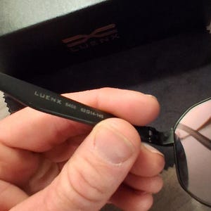 May include: Black framed sunglasses with pink lenses are held in a hand. The glasses are marked "LUENX 6406 52014-145". The glasses case is black with the words "SEE BETTER WORLD" in gold.