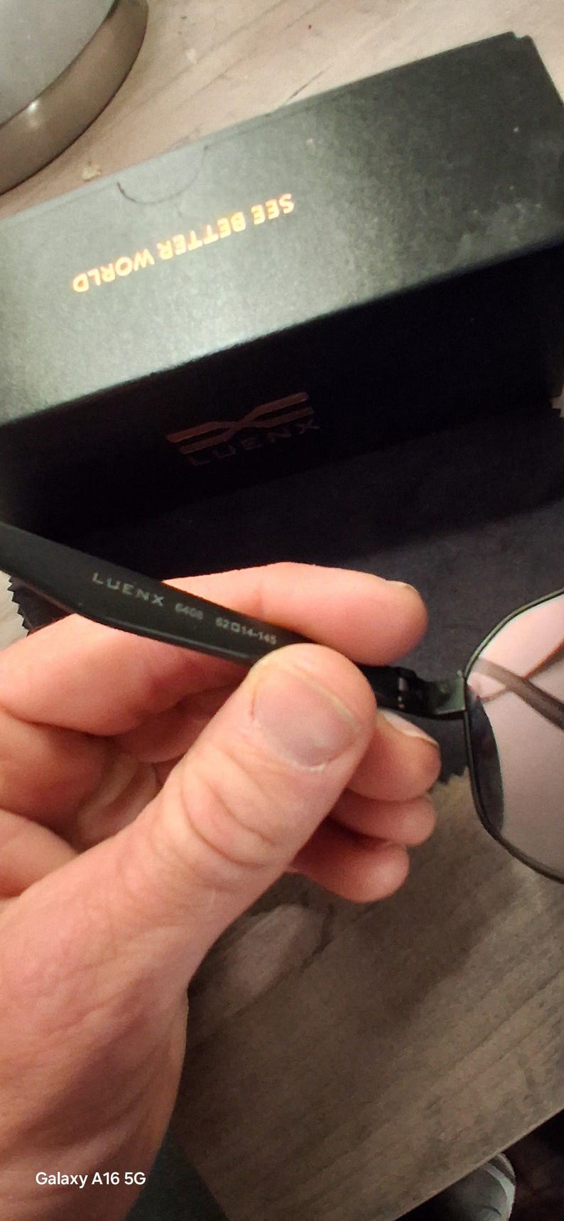 May include: Black framed sunglasses with pink lenses are held in a hand. The glasses are marked "LUENX 6406 52014-145". The glasses case is black with the words "SEE BETTER WORLD" in gold.