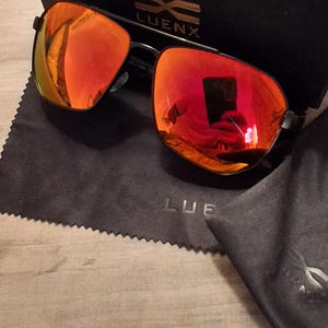 May include: Black framed sunglasses with mirrored orange lenses, a black case, and a black cleaning cloth. The brand name "LUENX" is visible on the case and cloth. The sunglasses reflect the surrounding environment.