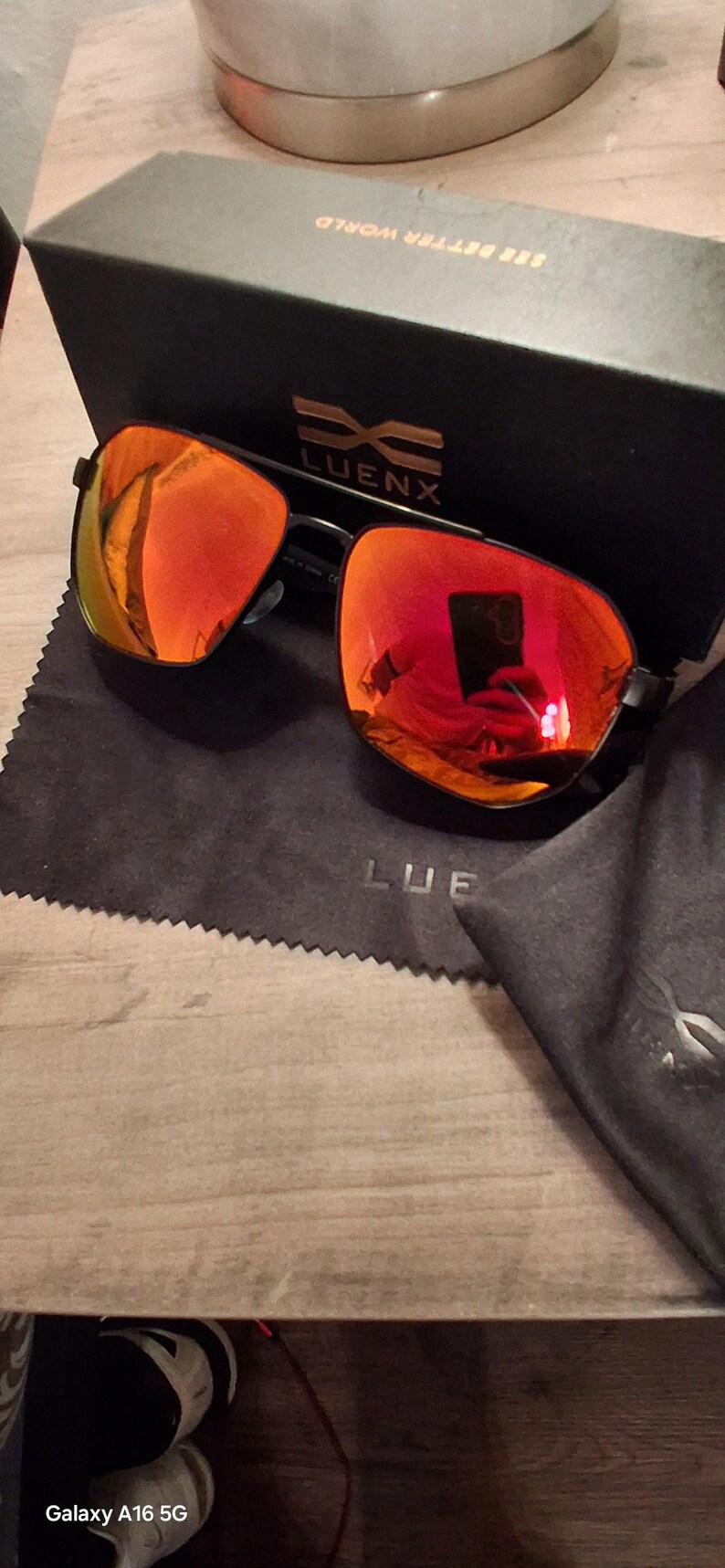 May include: Black framed sunglasses with mirrored orange lenses, a black case, and a black cleaning cloth. The brand name "LUENX" is visible on the case and cloth. The sunglasses reflect the surrounding environment.