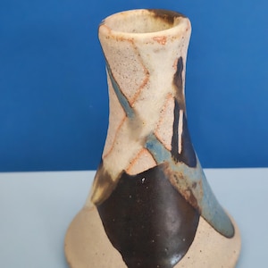 May include: A ceramic vase with a flared base and a narrow neck, featuring a speckled off-white surface decorated with abstract patches of dark brown, black, and muted blue glaze. The texture appears matte and granular throughout the exterior.