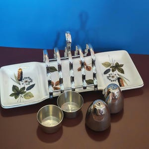 May include: A white ceramic toast rack with a central chrome metal handle and botanical illustrations of leaves and seed heads. The set includes two small round metal bowls and two matching dome-shaped metal salt and pepper shakers.