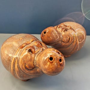 May include: Two brown ceramic hippopotamus-shaped shakers with a glossy, speckled glaze finish. One shaker features three small holes on the top of its head, while the other features a single hole, both designed for dispensing salt and pepper.