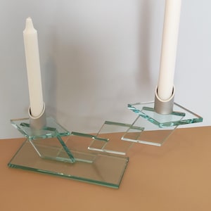 May include: A clear glass candle holder featuring a stepped, geometric design with two silver-toned cylindrical sockets. Two tall, slender white taper candles are inserted into the sockets, which are positioned at different heights on the glass structure.