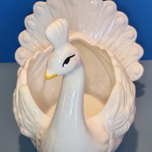 May include: A cream-coloured ceramic peacock planter featuring a glossy finish with a subtle crackle texture. The bird has a raised, fanned tail, a long neck, and a small yellow beak. The central body of the peacock forms a hollow basin.