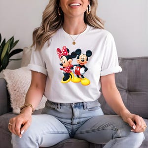 May include: White t-shirt featuring a colorful graphic of Mickey Mouse and Minnie Mouse. Minnie Mouse is wearing a red polka dot bow and dress. Mickey Mouse is wearing red shorts and yellow shoes. The shirt has rolled-up sleeves.