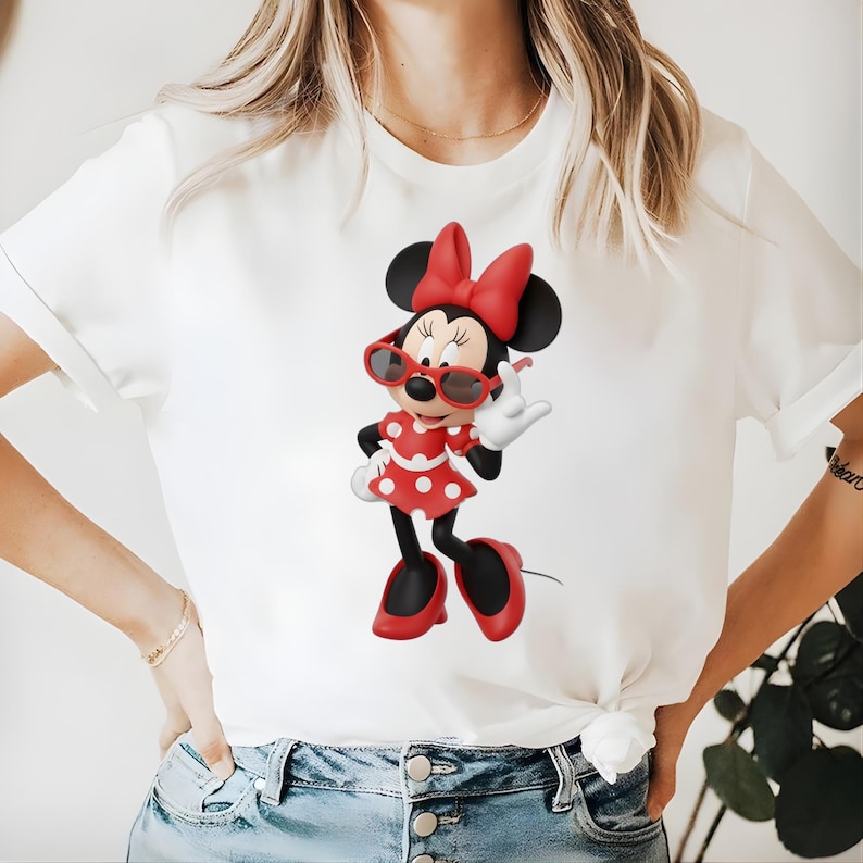 May include: White t-shirt featuring a cartoon Minnie Mouse character. Minnie Mouse is wearing red sunglasses, a red bow, and a red and white polka dot dress. The character is making a hand gesture.